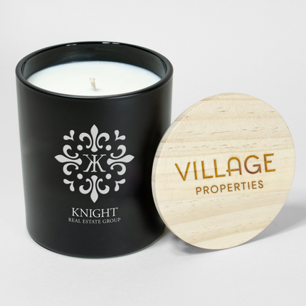 A 14 oz Deluxe "Design Your Own" Candle in a black glass vessel from Custom Candles LA. The candle features an etched logo that reads "Knight Real Estate Group" on the glass vessel and a wooden lid engraved with text that reads "Village Properties".  The candle if filled with white wax.