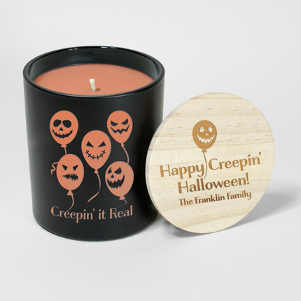 The Holiday "Creepin' It Real - Balloons" candle is a black candle from Custom Candles LA, featuring orange wax and a white wick. The vessel is etched with 5 balloons with jack o lantern faces with the text "creepin' it real" below; it comes next to a wooden lid engraved with a single jack o lantern balloon and the text "Happy Creepin' Halloween! The Franklin Family"