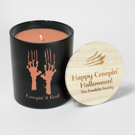 The Holiday "Creepin' It Real - Claws" candle is a black candle from Custom Candles LA, featuring orange wax and a white wick. The vessel is etched with two hands and claw marks with the text "creepin' it real" below; it comes next to a wooden lid engraved with claw marks and the text "Happy Creepin' Halloween! The Franklin Family"