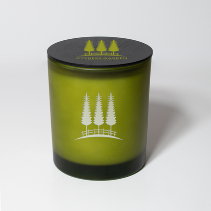 A 14 oz Deluxe "Design Your Own" Candle in a  frosted olive glass vessel from Custom Candles LA. The candle features an etched logo of cypress tress on the glass vessel with white wax showing through and a black wooden lid imprinted with a logo of cypress trees that reads "Cypress Garden Creative Design" in green.