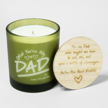 A **green Glad You're My Dad Candle — Premium by Custom Candles LA** with the text "Glad You're My DAD" and "Love Elizabeth" on the side. The wooden lid, placed next to the candle, features custom engravings: "For my Dad, who taught me how to sail, ski, and open a bottle of champagne. You're the Best EVER!" A truly premium gift.