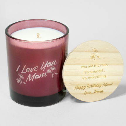 The "I Love You Mom" candle is in a wine vessel from Custom Candles LA, featuring white wax and a white wick. The candle is etched with the text "I Love You Mom" in a script font with decorative flowers surrounding; it comes next to a wooden lid engraved with the text "You are my rock, my strength, my everything. happy Birthday Mom! Love, Jenna" and floral design above the text.