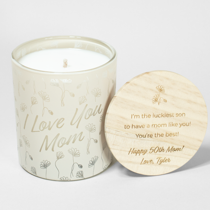 The "I Love You Mom" candle is in a cream vessel from Custom Candles LA, featuring white wax and a white wick. The candle is etched with the text "I Love You Mom" in a script font with decorative flowers surrounding; it comes next to a wooden lid engraved with the text "I'm the luckiest son to have a mom like you! You're the best! Happy 50th Mom! Love, Tyler" and floral design above the text.