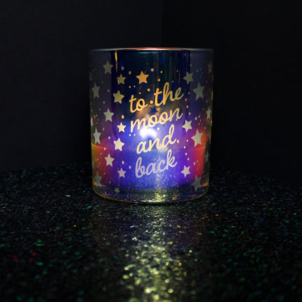 The "To the Moon and Back Anniversary" candle is featured in an iridescent candle from Custom Candles LA, with the text "to the moon and back" etched into the glass with stars and dots surrounding the text. The candle is lit and is sitting on a sparkly black floor with a plain black background.