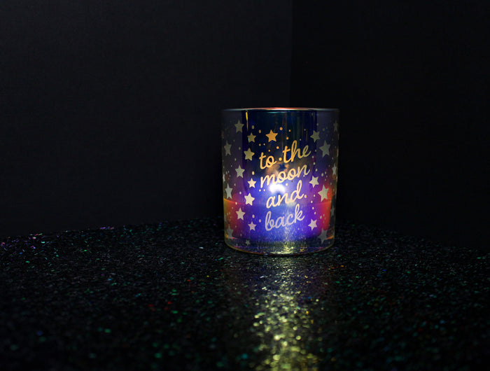 The "To the Moon and Back Anniversary" candle is featured in an iridescent candle from Custom Candles LA, with the text "to the moon and back" etched into the glass with stars and dots surrounding the text. The candle is lit and is sitting on a sparkly black floor with a plain black background.