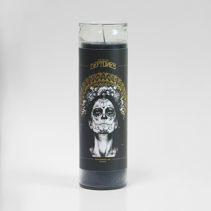 An unscented 16 oz prayer candle from Custom Candles LA. The candle is encased in a tall, clear glass vessel with a black wax interior. The front of the candle features an intricate design with a white, skull-painted face resembling a Day of the Dead motif, surrounded by a gold ornate pattern. Above the face, the text "DIA DE LOS DEFTONES" is written in gold. At the bottom, it reads "SAN DIEGO, CA 2022." The candle is unlit, with a single wick visible. The design is striking and artistic.