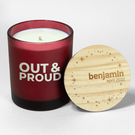 This "Out & Proud" candle is featured  in a frosted red glass from Custom Candles L.A., labeled "OUT & PROUD" on the glass with white wax showing through. The wooden listar designs.d is engraved with "benjamin" and "april 2022," along with illustrative sparkles and stars surrounding the inside of the circular lid.