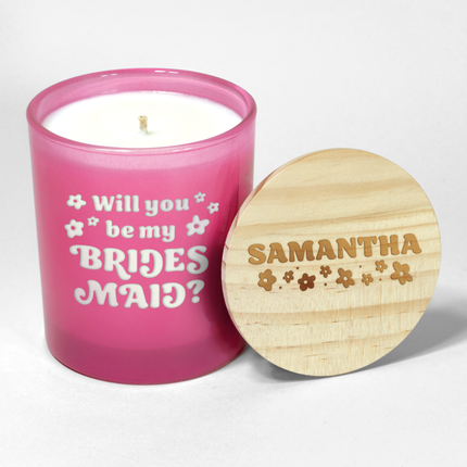 The "Will you be my Bridesmaid?" candle is a hot pink candle from Custom Candles LA, featuring white wax and a white wick and labeled "Will you be my Bridesmaid?" in a funky font with decorative flowers surrounding; it comes next to a wooden lid engraved with the name "Samantha" and floral designs, making it an ideal wedding party gift.