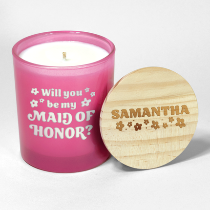 The "Will you be my Bridesmaid?" candle is a hot pink candle from Custom Candles LA, featuring white wax and a white wick and labeled "Will you be my Maid of Honor?" in a funky font with decorative flowers surrounding; it comes next to a wooden lid engraved with the name "Samantha" and floral designs, making it an ideal wedding party gift.