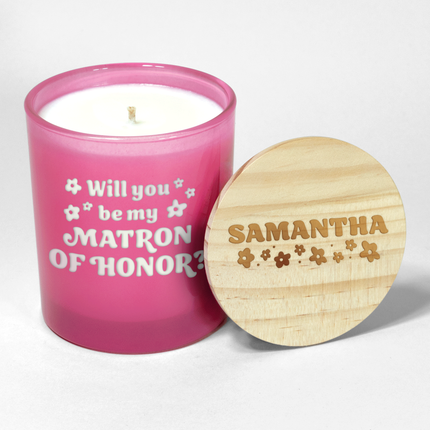 The "Will you be my Bridesmaid?" candle is a hot pink candle from Custom Candles LA, featuring white wax and a white wick and labeled "Will you be my Matron of Honor?" in a funky font with decorative flowers surrounding; it comes next to a wooden lid engraved with the name "Samantha" and floral designs, making it an ideal wedding party gift.