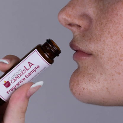 Person smelling a Custom Candles LA fragrance sample bottle close to their nose against a gray background