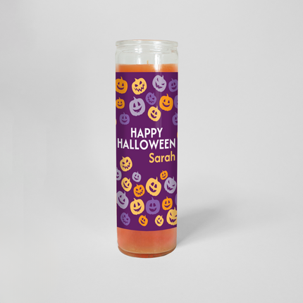 This image shows a 16 oz prayer candle from Custom Candles LA. The candle, filled with orange wax, features a purple label printed with multicolored jack-o-lanters and the text "Happy Halloween Sarah" printed in the middle.