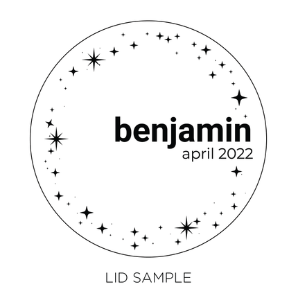 A sample of the wooden lid for the "Out & Proud" by Custom Candles L.A. features the name "Benjamin" and date "April 2022"across the middle surrounded by illustrative sparkles surrounding the edge of the circular lid. 