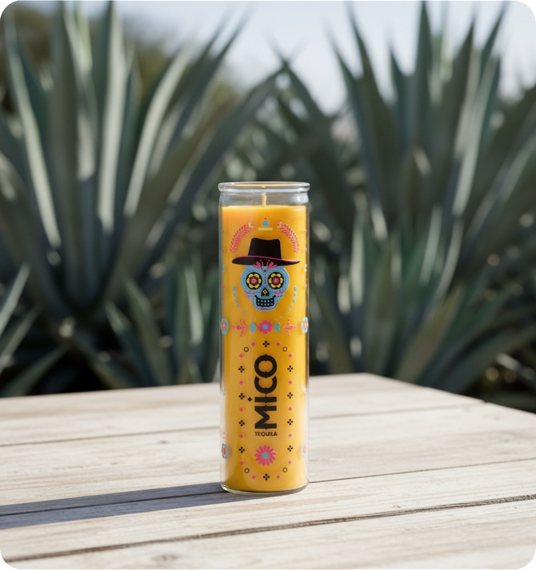 a yellow wax prayer candle wtih a colorful design imprint set down on an ash white wooden tabletop, surrounded by several natural agave plants.