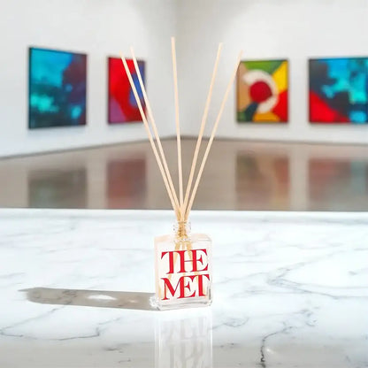 2.5 oz clear Reed diffuser with branding on a marble surface with colorful abstract paintings in the background.