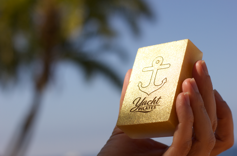 Hand holding a gold rectangle bar soap engraved with an anchor logo against palm trees