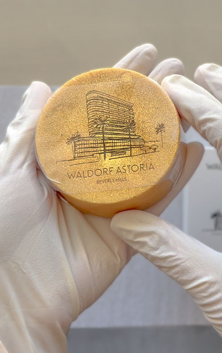 Gloved hands holding a gold round bar soap engraved with a building illustration