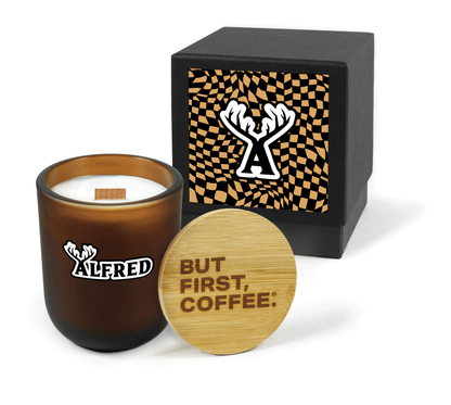 A 12 oz Luxe Custom Candle in the color “Cacao” which is an amber brown from Custom Candles LA. The candle features a wood wick and a black and white imprinted logo for “Alfred Coffee” on the front of the glass. Leaning against the side of the candle is a bamboo candle lid etched with “But First, Coffee®“. In the background, there is a black 2-piece box 4-color printed with an abstract tan and black square pattern with the icon logo for “Alfred Coffee” featuring an A with horns.