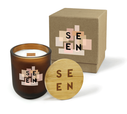 A 12 oz Luxe Custom Candle in the color “Cacao” which is an amber brown from Custom Candles LA. The candle features a wood wick and 4-color printed logo for “SEEN” on top of various square in shades of pink on the front of the glass. Leaning against the side of the candle is a bamboo candle lid etched with the “Seen” logo. In the background, there is a kraft 2-piece box 4-color printed with the “Seen” logo on top of squares in shades of pink.