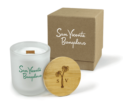 
A 12 oz Luxe Custom Candle in the color “Cashmere” which is a diffused white from Custom Candles LA. The candle features a wood wick and 4-color green printed logo for “San Vicente Bungalows” on the front of the glass. Leaning against the side of the candle is a bamboo candle lid etched with a palm tree and “S” and “V”. In the background, there is a kraft 2-piece box printed with a white logo for “San Vicente Bungalows”.