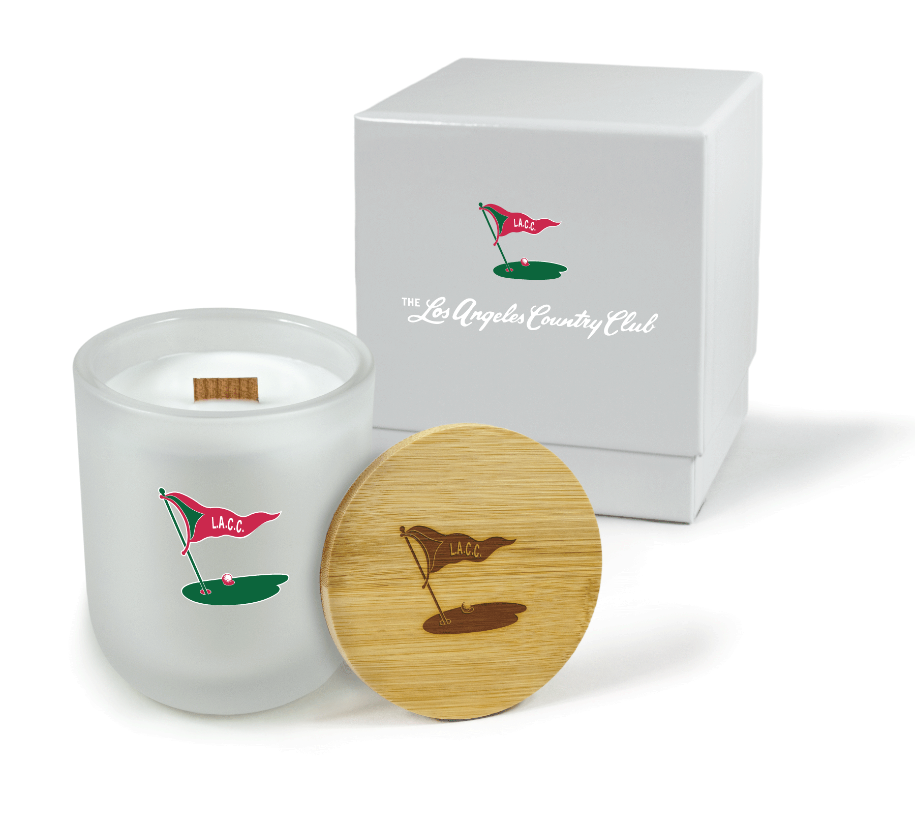 A 12 oz Luxe Custom Candle in the color “Cashmere” which is a diffused white from Custom Candles LA. The candle features a wood wick and pink and green icon logo for “Los Angeles Country Club” made up of a golf green, ball and flag reading “L.A.C.C.” on the front of the glass. Leaning on the candle is a bamboo candle lid etched with the same logo icon. In the background, there is a white 2-piece box printed with a white logo for “The Los Angeles Country Club” below the 4-color L.A.C.C. icon logo.