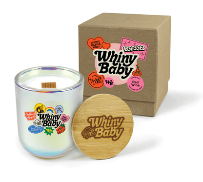 A 12 oz Luxe Custom Candle in the color “Bliss” which is an iridescent pink from Custom Candles LA. The candle features a wood wick and 4-color printed logo for “Whiny Baby” surrounded by decorative sticker style graphics on the front of the glass. Leaning against the side of the candle is a bamboo candle lid etched with the “Whiny Baby” logo. In the background, there is a kraft 2-piece box 4-color printed with a white and black “Whiny Baby” logo surrounded by pink and red sticker style graphics.