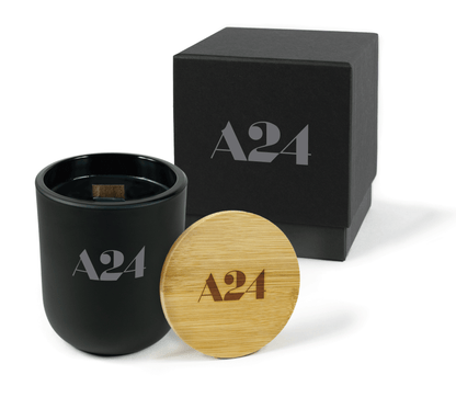 A 12 oz Luxe Custom Candle in the color “Darkest Night” which is jet black from Custom Candles LA. The candle features a wood wick and gray 4-color imprinted logo for “A24" on the front of the glass. Leaning against the side of the candle is a bamboo candle lid etched with the “A24” logo. In the background, there is a black 2-piece box 4-color printed with a grey “A24" logo.