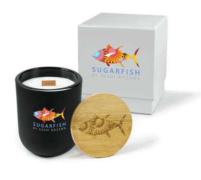 A 12 oz Luxe Custom Candle in the color “Darkest Night” which is a jet black from Custom Candles LA. The candle features a wood wick and a 4-color “Sugarfish by Sushi Nozawa” logo below a multi-colored and patterned fish logo on the front of the glass. Leaning against the side of the candle is a bamboo candle lid etched with the the multi-patterned fish logo. In the background, there is a white 2-piece box 4-color printed with the same “Sugarfish by Sushi Nozawa” and colorful fish logos.