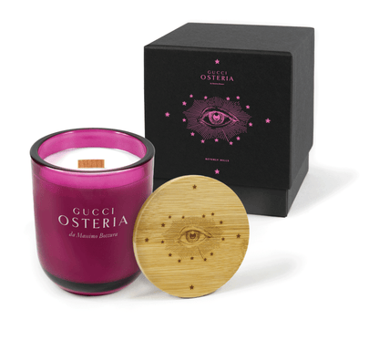 A 12 oz Luxe Custom Candle in the color “Cherry Kiss” which is a juicy magenta from Custom Candles LA. The candle features a wood wick and logo for “Gucci Osteria da Massimo Bottura” engraved on the front of the glass showing the white wax of the candle. Leaning against the side of the candle is a bamboo candle lid etched with an intricate eye logo surrounded by lines and stars. In the background, there is a black 2-piece box 4-color printed with both of those logos in bright pink.