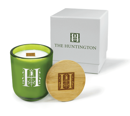 A 12 oz Luxe Custom Candle in the color “Grassland” which is a verdant green from Custom Candles LA. The candle features a wood wick and an engraved logo symbol of a decorated H for “The Huntington” on the front of the glass. Leaning against the side of the candle is a bamboo candle lid etched with same decorative H icon. In the background, there is a white 2-piece box, 4-color printed with a green and white logo for "The Huntington".