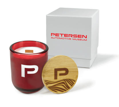 A 12 oz Luxe Custom Candle in the color “Blood Red” which is a vivid rouge red from Custom Candles LA. The candle features a wood wick and an engraved “P” on the front of the glass showing the white wax of the candle. Leaning against the side of the candle is a bamboo candle lid etched with the a “P” and an abstract mountain pattern. In the background, there is a white 2-piece box printed with red logo for “Petersen Automotive Museum”.