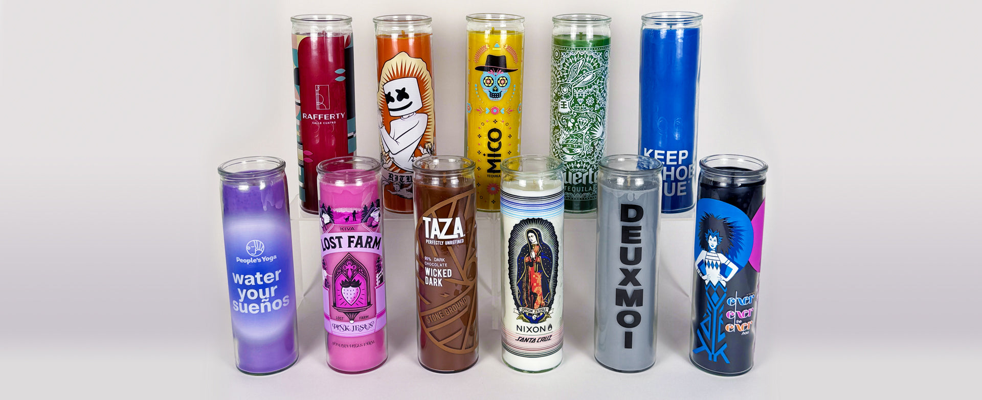 a wide group shot of 11 prayer candles all with different color waxes and different logos and designs on each one, arranged in two tiers with 6 being in front and 5 being behind on an elevated platform