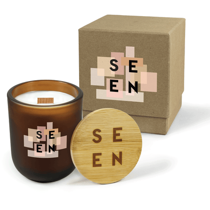 A 12 oz Luxe Custom Candle in the color “Cacao” which is an amber brown from Custom Candles LA. The candle features a wood wick and 4-color printed logo for “SEEN” on top of various square in shades of pink on the front of the glass. Leaning against the side of the candle is a bamboo candle lid etched with the “Seen” logo. In the background, there is a kraft 2-piece box 4-color printed with the “Seen” logo on top of squares in shades of pink.