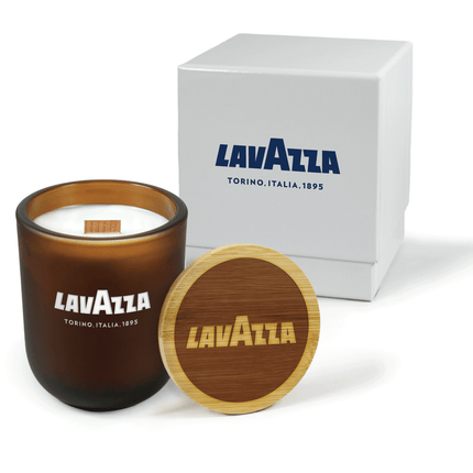 A 12 oz Luxe Custom Candle in the color “Cacao” which is an amber brown from Custom Candles LA. The candle features a wood wick and “Lavazza Torina. Italia. 1895” engraved on the front of the glass showing the white wax of the candle. Leaning against the side of the candle is a bamboo candle lid etched with the the “Lavazza” logo. In the background, there is a white 2-piece box printed with blue logo for “Lavazza” above text reading “Torina. Italia. 1895".