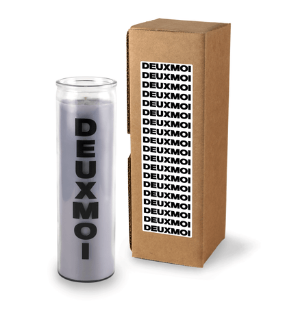 A 16 oz Prayer Candle from Custom Candles LA filled with gray wax in a tall glass container. The candle is printed with a logo that reads "DEUXMOI" in bold black letters that are stacked vertically down the front of the glass. To the right of the candle sits a Kraft mailer box with a sticker on the box that is printed with "DEUXMOI" in bold black letters that repeats itself in a stacking pattern  on a white background. 