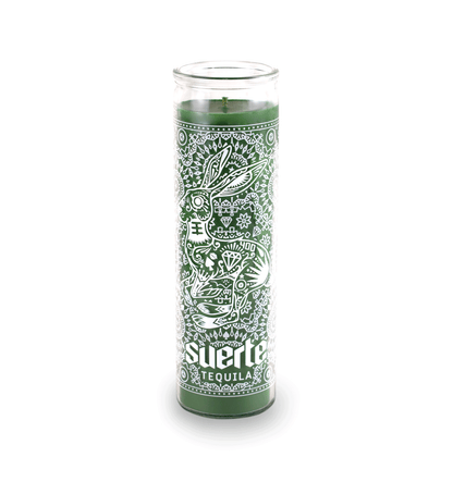 A 16 oz Prayer Candle from Custom Candles LA filled with green wax in a tall glass container. The candle is printed a logo at the bottom that reads "Suerte Tequila" in white. The logo is surrounded by an ornate pattern that makes the shape of a rabbit. The pattern includes diamonds, dots, flowers, and other line artwork. The green wax shows through the artwork.