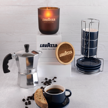 A lit 12 oz Luxe Candle in "Cacao" an amber brown color from Custom Candles L.A with . The candle is etched with a logo that reads "Lavazza" with the flame glowing through. It sits on a white box with the Lavazza logo printed in blue.  Leaning on the right side of the box is a wooden candle lid that is engraved with the Lavazza logo in a circle. All three items are surrounded with a coffee press, coffee beans, a mug of coffee with a cookie, and more mugs. The foreground and background are both white.
