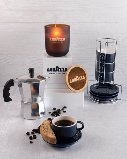 A lit 12 oz Luxe Candle in "Cacao" an amber brown color from Custom Candles L.A with . The candle is etched with a logo that reads "Lavazza" with the flame glowing through. It sits on a white box with the Lavazza logo printed in blue.  Leaning on the right side of the box is a wooden candle lid that is engraved with the Lavazza logo in a circle. All three items are surrounded with a coffee press, coffee beans, a mug of coffee with a cookie, and more mugs. The foreground and background are both white.