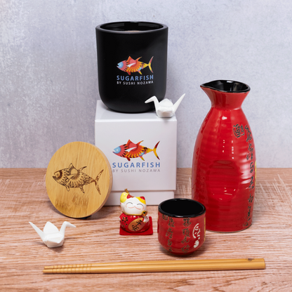 A 12 oz Luxe Candle in "Darkest Night" a jet black color from Custom Candles L.A. The candle is printed with a colorful logo of "sugarfish by sushi nozawa". It sits next to a white crane and both sit on a white box printed with the same colorful logo.  Leaning on the left side of the box is a wooden candle lid that is engraved with the fish from the logo. In the foreground sits a red sake set and a pair of wooden chopsticks. Everything sits on a wooden table and a gray background. 