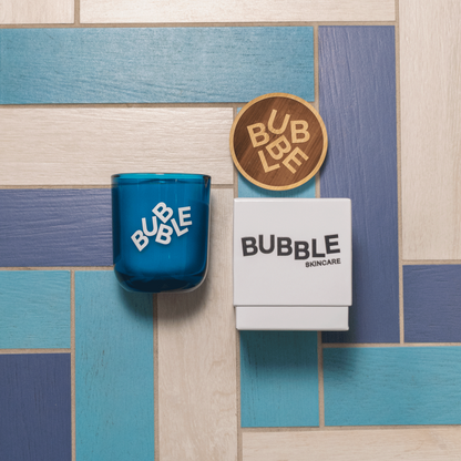 A 12 oz Luxe Candle in "Oceania" a splashy teal color from Custom Candles L.A. The candle is printed with a logo that reads "BUBBLE" in white scrambled letters. On the right sits a white box with the logo that reads "BUBBLE SKINCARE" printed in black.  Above the box is a wooden lid that is engraved with the "BUBBLE" logo in scrambled letters. It is a top down view, and the background has blue, navy, and cream colored rectangular tiles.
