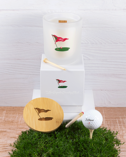 A 12 oz Luxe Candle in "Cashmere" a diffused white from Custom Candles L.A with white wax and a wooden wick. The candle is printed with the "L.A.C.C." logo in red & green of a golf flag, ball, and green. It sits next to a white tee on a white box with same logo and "Los Angeles Country Club printed.  Leaning on the left side of the box is a wooden candle lid that is engraved with the same logo. They sit on a patch of grass next to a pencil and a teed up golf ball on a wooden floor and white wood background.