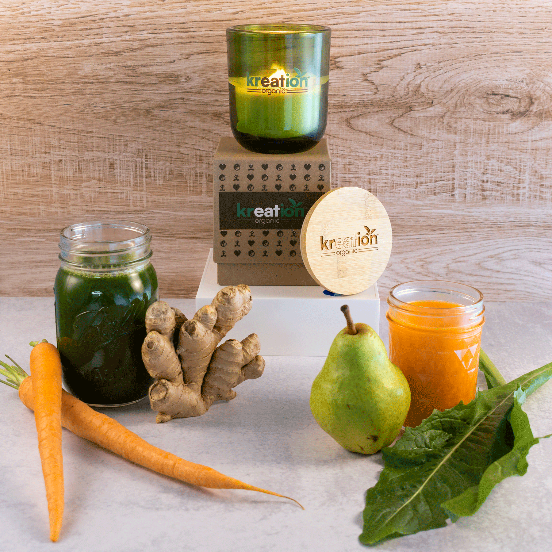 A lit 12 oz Luxe Candle in "Grassland" a verdant green color from Custom Candles L.A. The candle is printed with a logo that reads "kreation organic" in green and brown. It sits on a kraft box with the same logo printed on it surrounded by various icons.  Leaning on the right side of the box is a wooden candle lid that is engraved with the same logo. All three items sit on a white box. In the foreground there's ginger, carrots, juices, pear, and lettuce. They sit on a white table with a wooden background.