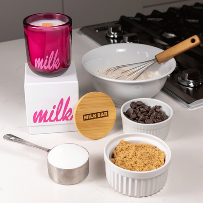 A 12 oz Luxe Candle in "Cherry Kiss" a juicy magenta color from Custom Candles L.A with white wax and a wooden wick. The candle is printed with a logo that reads "milk" in pink. It sits on a white box with the same logo printed in pink.  Leaning on the right side of the box is a wooden candle lid that is engraved with a logo that reads "Milk Bar" in a shaded rectangle. They sit on a white countertop next to a stove, surrounded by a bowl of flour with a whisk, chocolate chips, brown sugar, and white sugar.