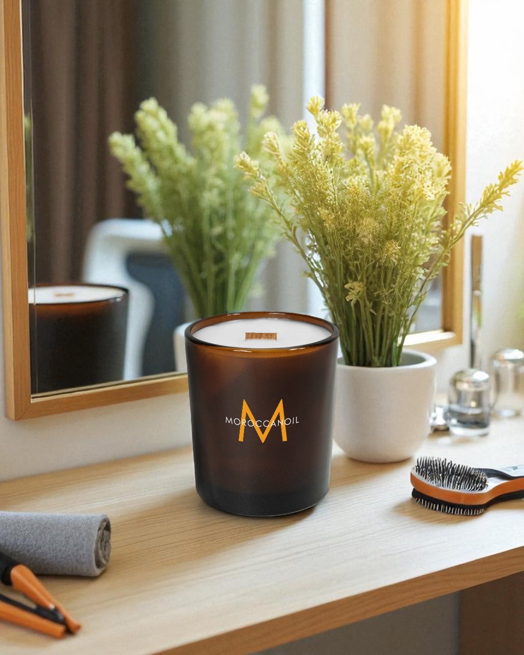 A amber 14 oz. glass candle with wooden wick featuring a colorful logo sits on a bamboo counter top next to a bright green plant  and other bathroom essentials in front of a mirror 