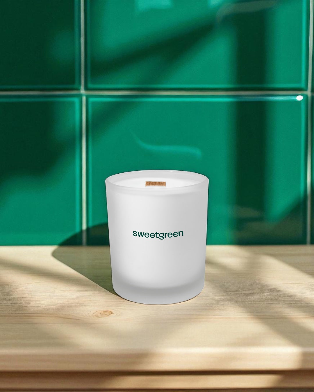 A frosted 14 oz. glass candle with wooden wick and green text sits on a light wood surface against a vibrant green tiled wall background