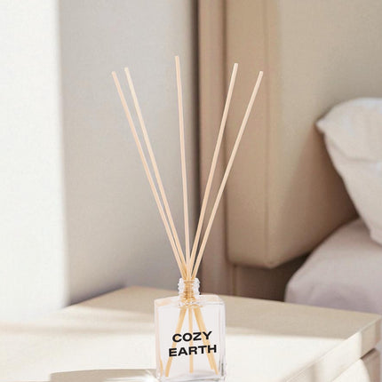 A clear glass reed diffuser with black text and wooden reed sticks sits on a white coffee table in a modern living room, with beige upholstered furniture and neutral decor in the background