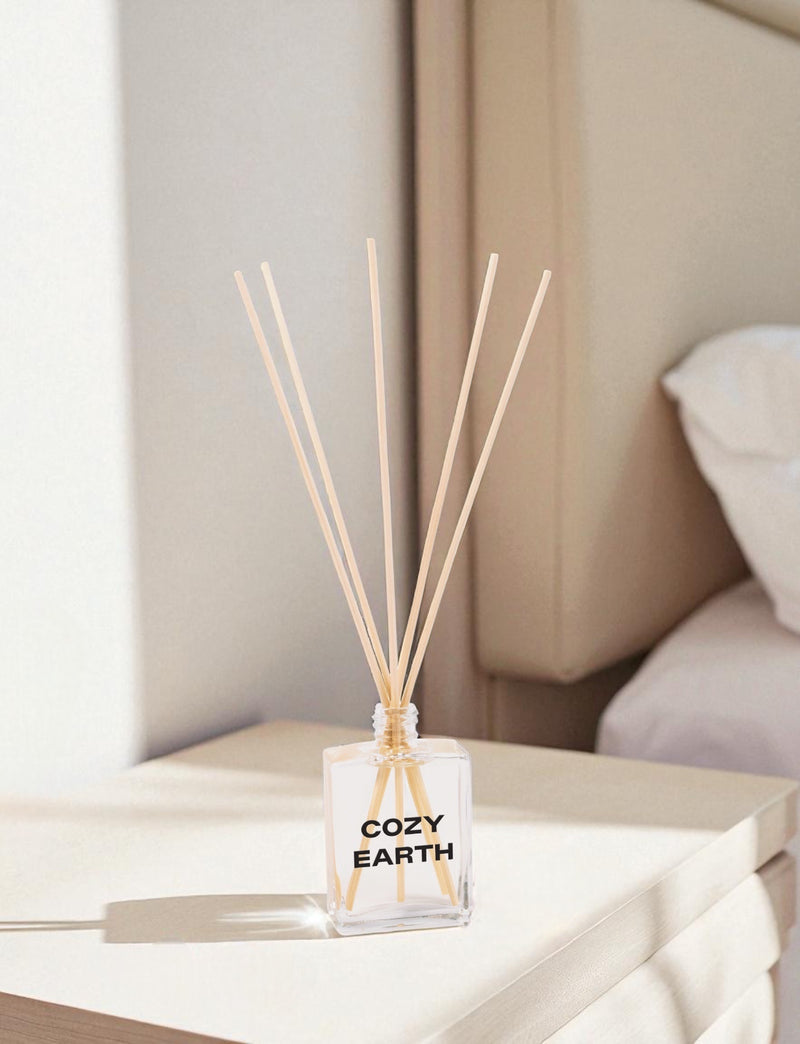 A clear glass reed diffuser with black text and wooden reed sticks sits on a white coffee table in a modern living room, with beige upholstered furniture and neutral decor in the background