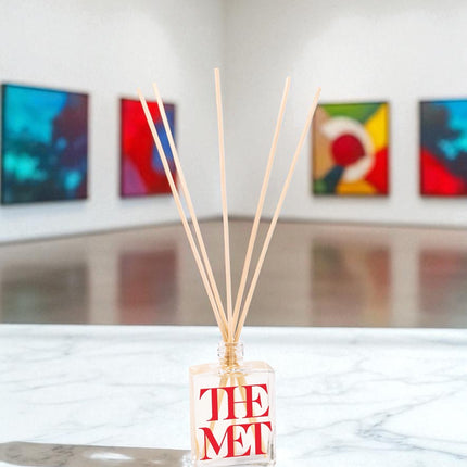 A clear glass reed diffuser with red text and wooden reed sticks sits on a white marble surface in an art gallery, with colorful abstract paintings displayed on white walls in the background