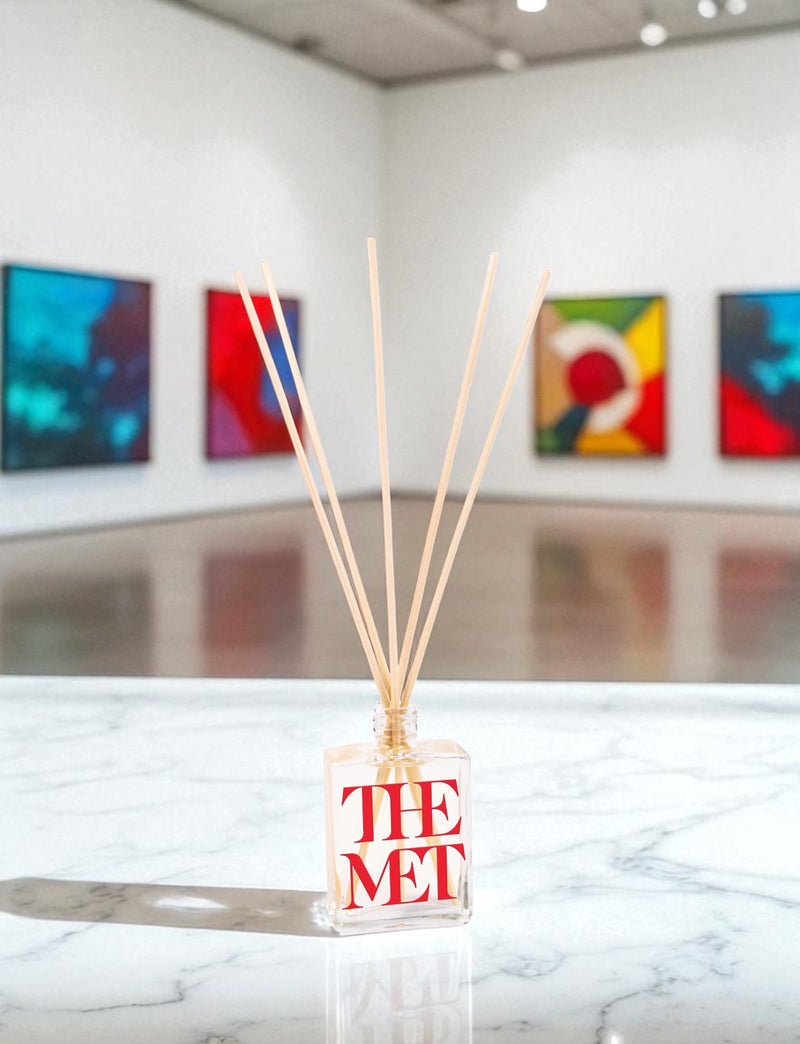 A clear glass reed diffuser with red text and wooden reed sticks sits on a white marble surface in an art gallery, with colorful abstract paintings displayed on white walls in the background