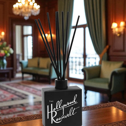 A black reed diffuser with white script text and black reed sticks sits on an ornate carved wooden coffee table in an elegant hotel lobby, with crystal chandeliers, rich wood paneling, green velvet curtains, and classic furnishings in the background